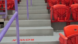 Stadium seating area featuring gray steps with a purple railing and red plastic seats, labeled with row numbers for easy navigation.